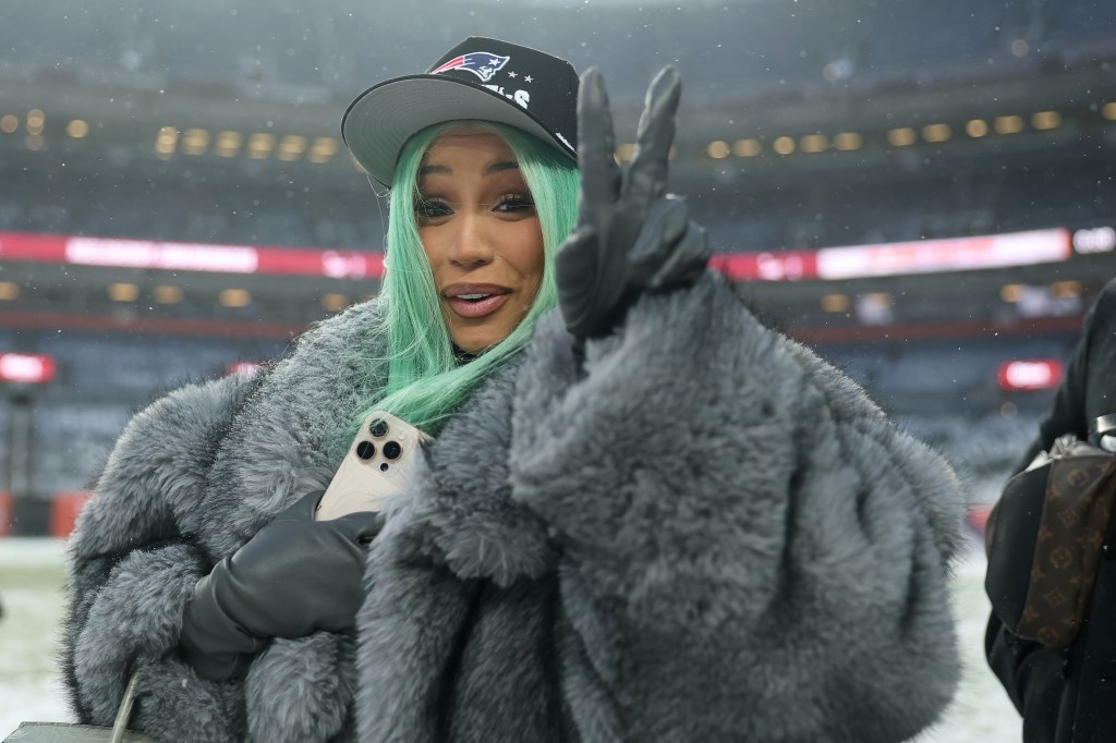 Cardi B wearing a New England Patriots cap and grey fur coat, giving a peace sign at an NFL game.