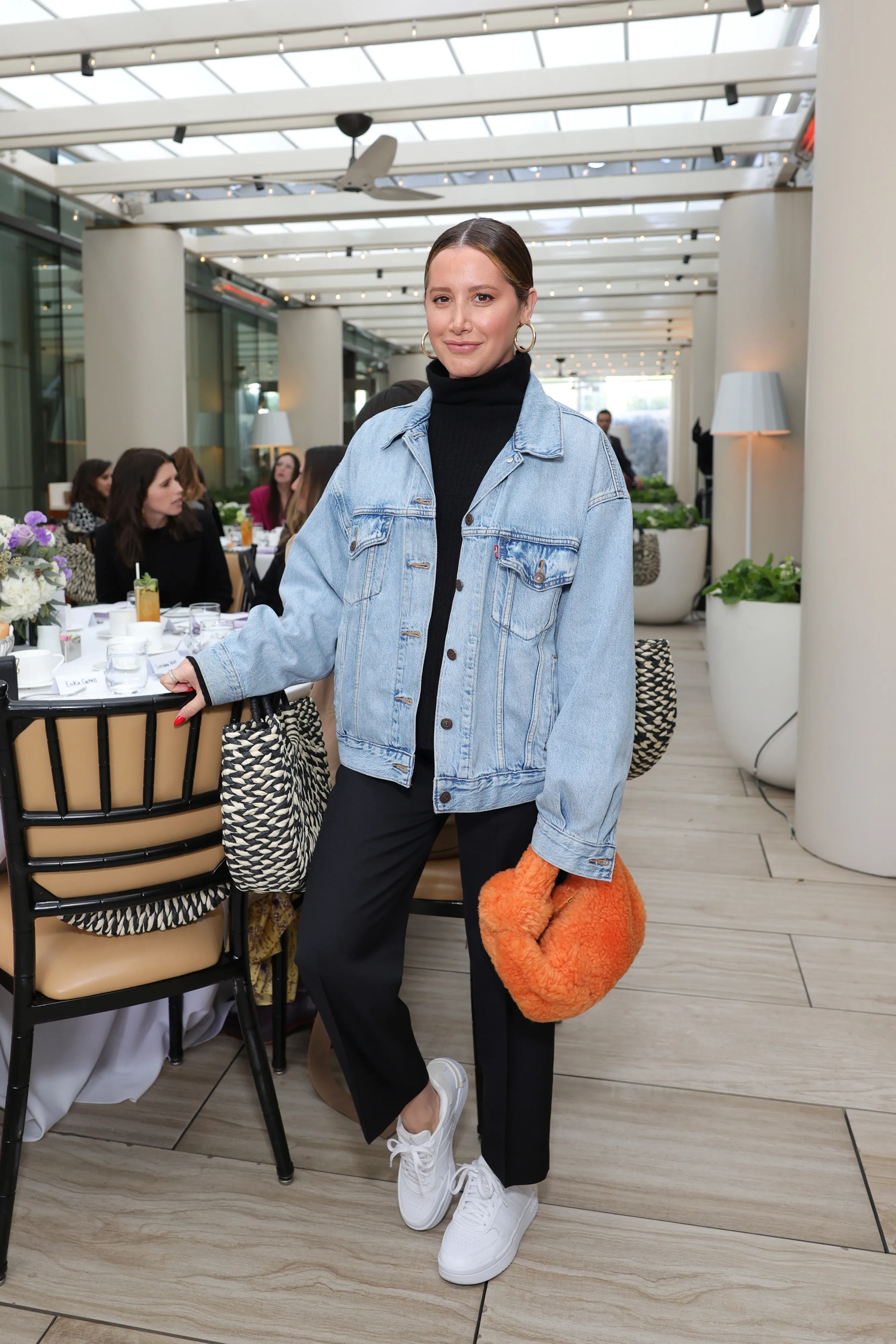 Ashley Tisdale standing and posing in a black turtleneck, denim jacket, black pants, white sneakers, and an orange fuzzy handbag.