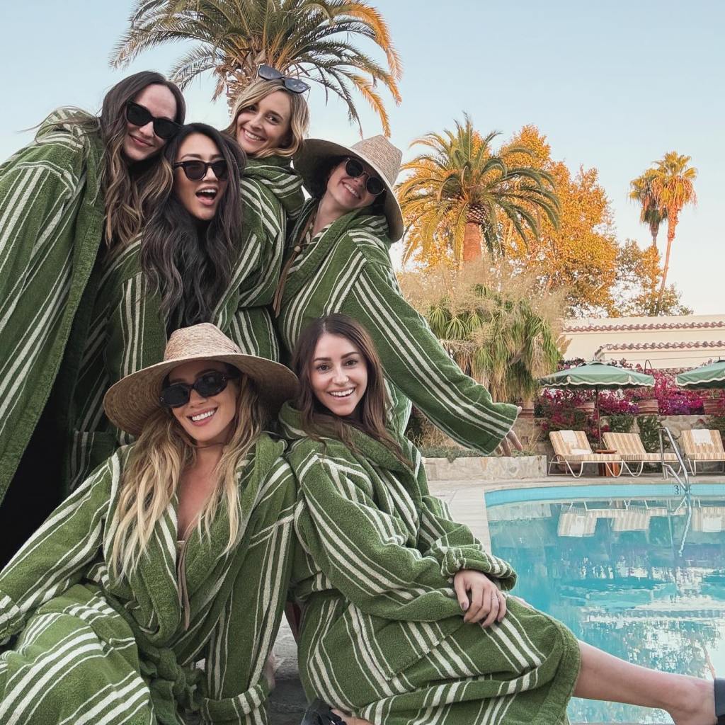 Hilary Duff and her mom group pose in green and white striped robes posing for a photo outdoors near a pool with palm trees.