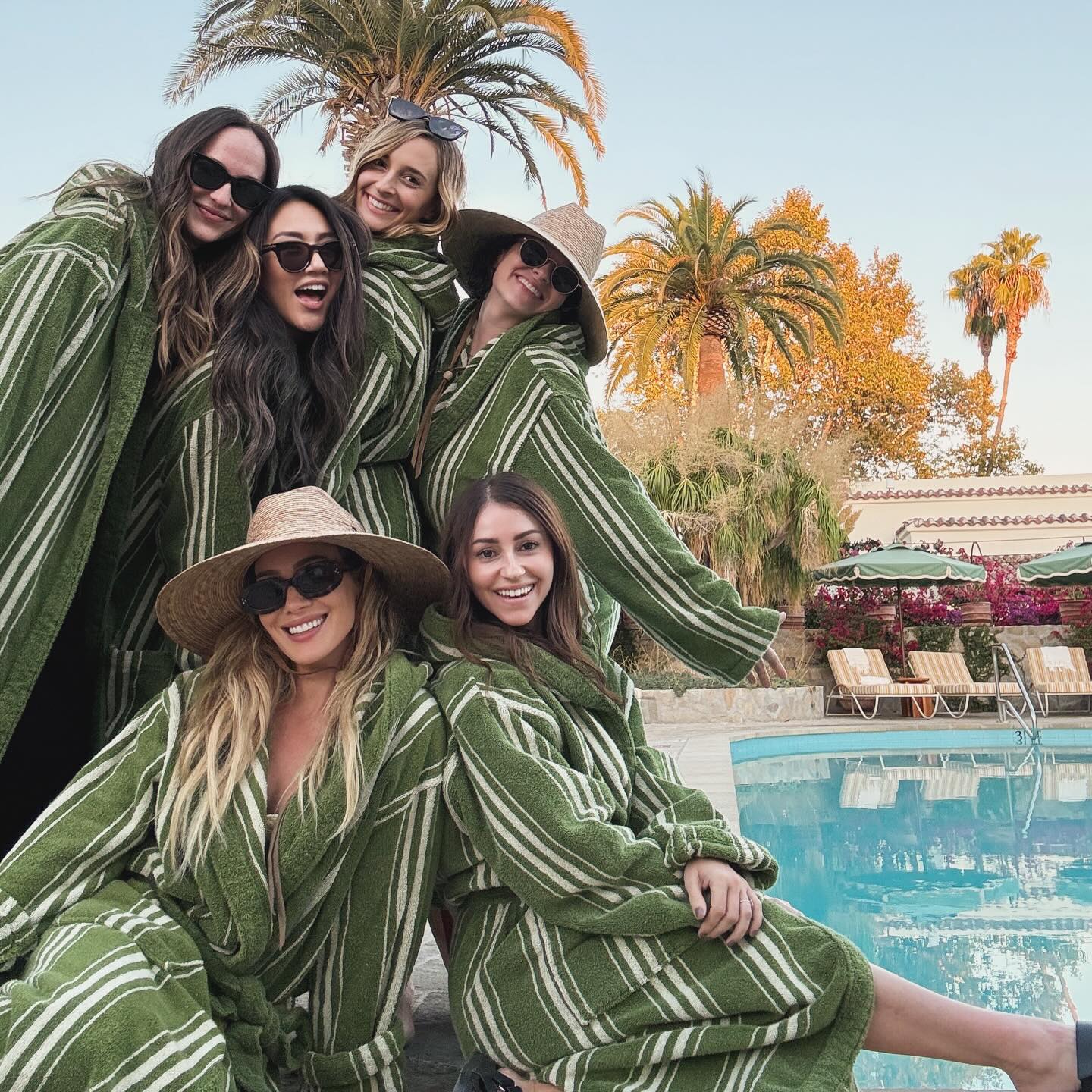 Six women in green and white striped robes by a pool with palm trees.