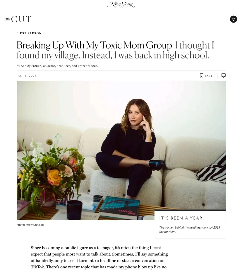 Ashley Tisdale sits on a cream-colored couch with a vase of flowers on a coffee table beside her.