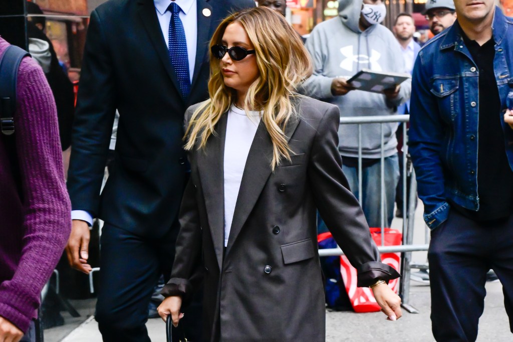 Ashley Tisdale in a long gray blazer, white shirt, and black cat-eye sunglasses, walking next to a man in a suit.