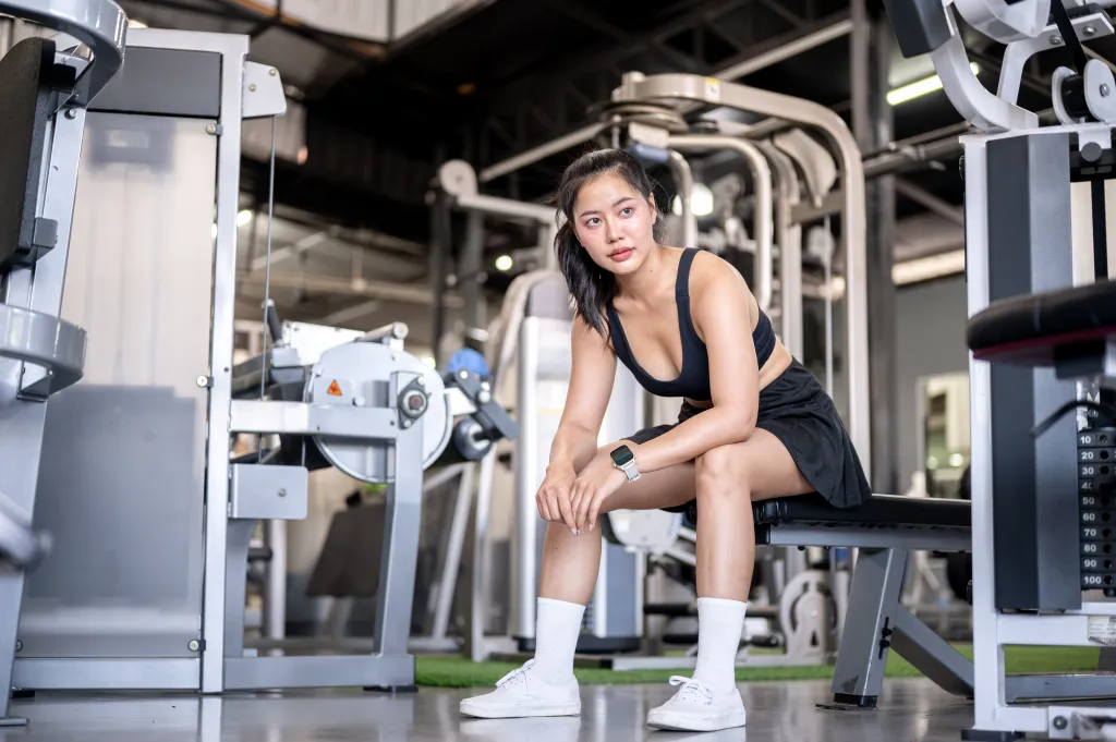 Asian woman in black sportswear sitting on a bench in a gym.