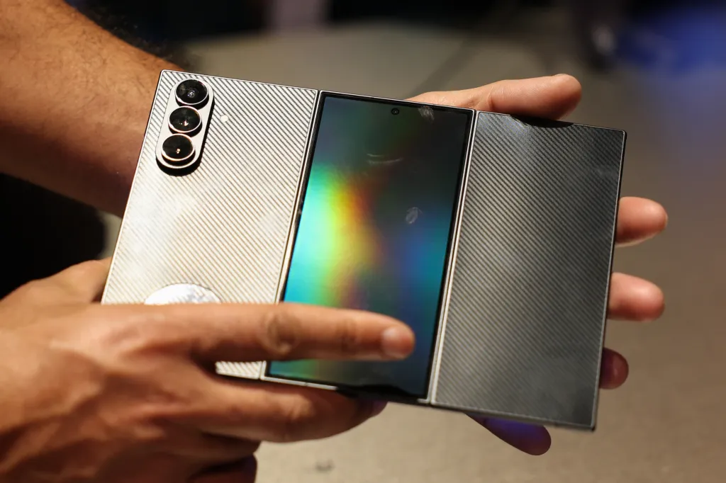 Hands holding a partially folded Samsung Galaxy Z Tri-Fold smartphone with a black, textured back and a screen displaying a rainbow spectrum.