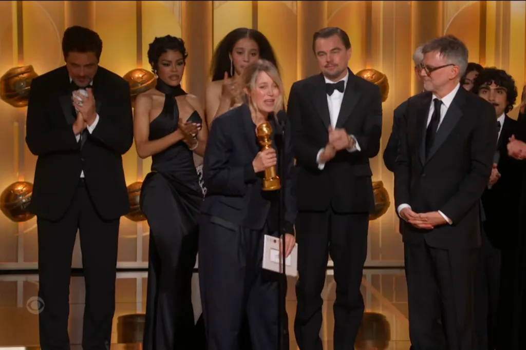 A woman in a dark pantsuit holds a Golden Globe award and speaks into a microphone, surrounded by other attendees in formal wear who are applauding her.