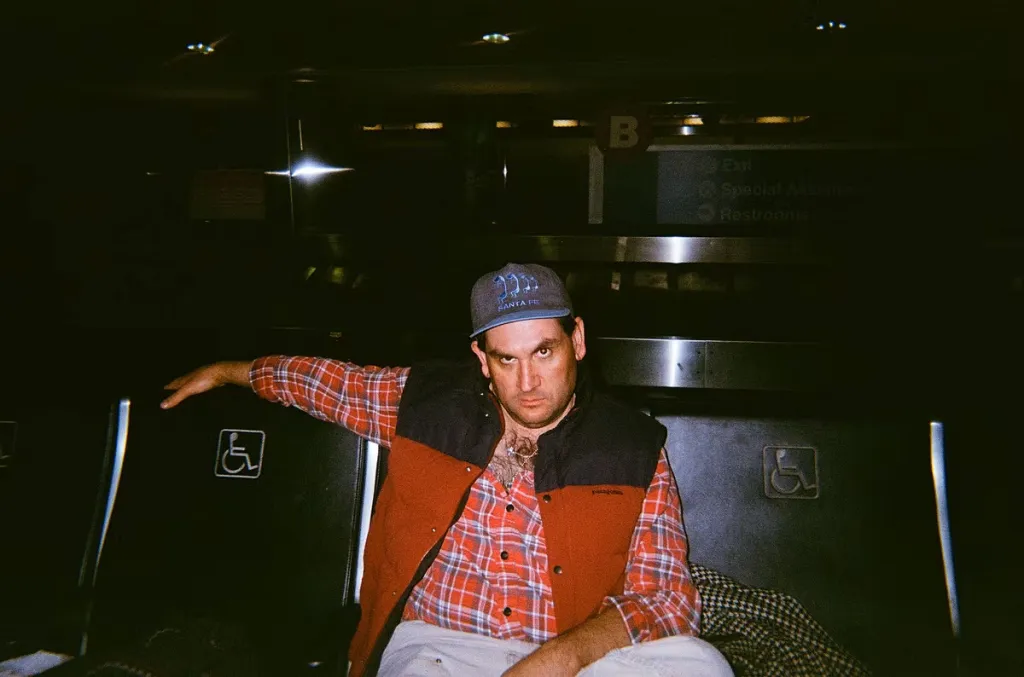 Austin Leonard Jones, son of Tommy Lee Jones, wearing a red and black vest over a red plaid shirt, a Santa Fe cap, and light pants, seated in a dimly lit public space with a dark background.