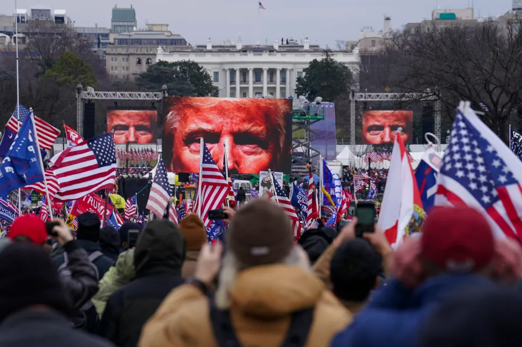 A crowd of Trump supporters carrying U.S. flags watch giant screens showing closeups on Trump's face.