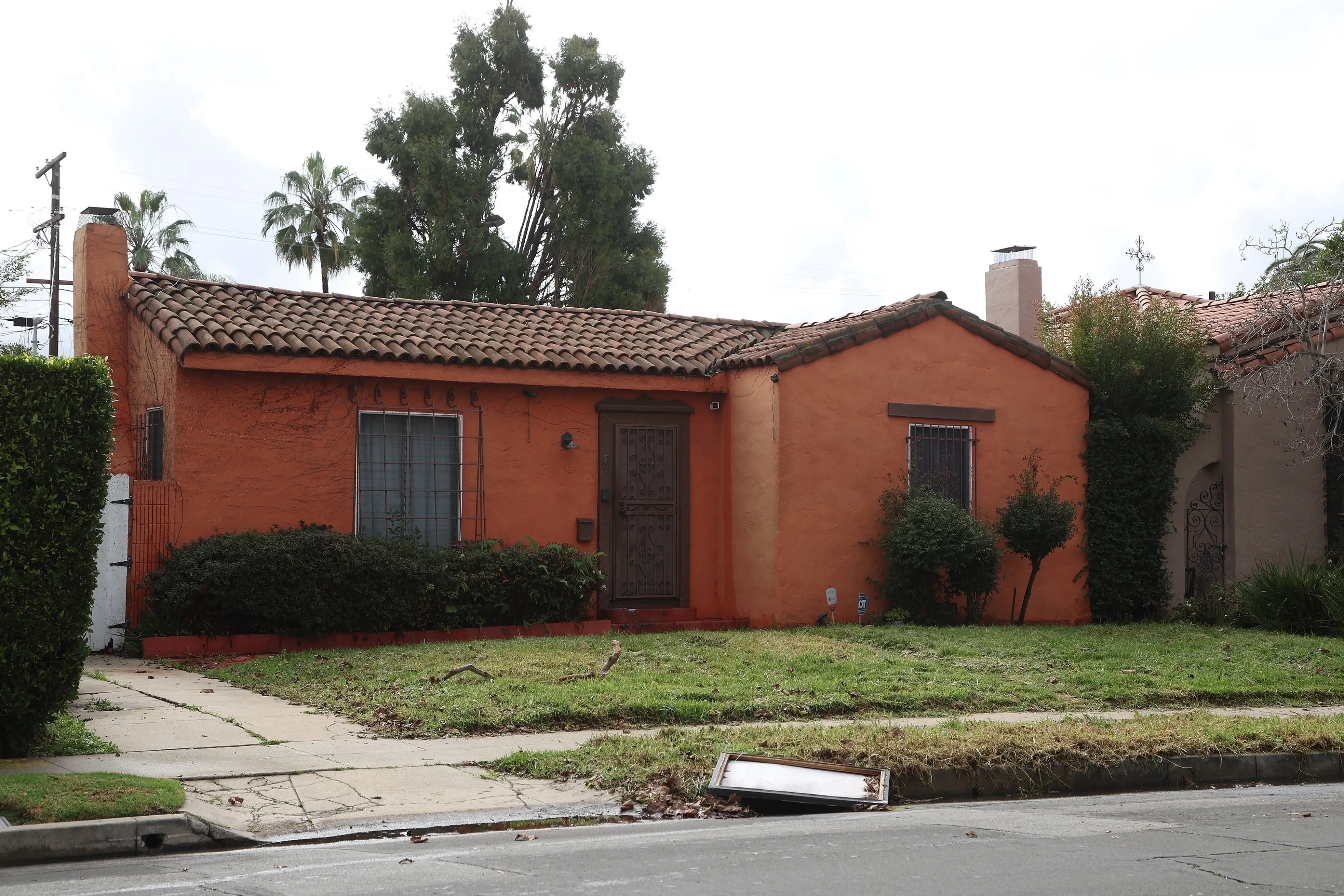 A small, orange stucco house with a tile roof and overgrown landscaping.