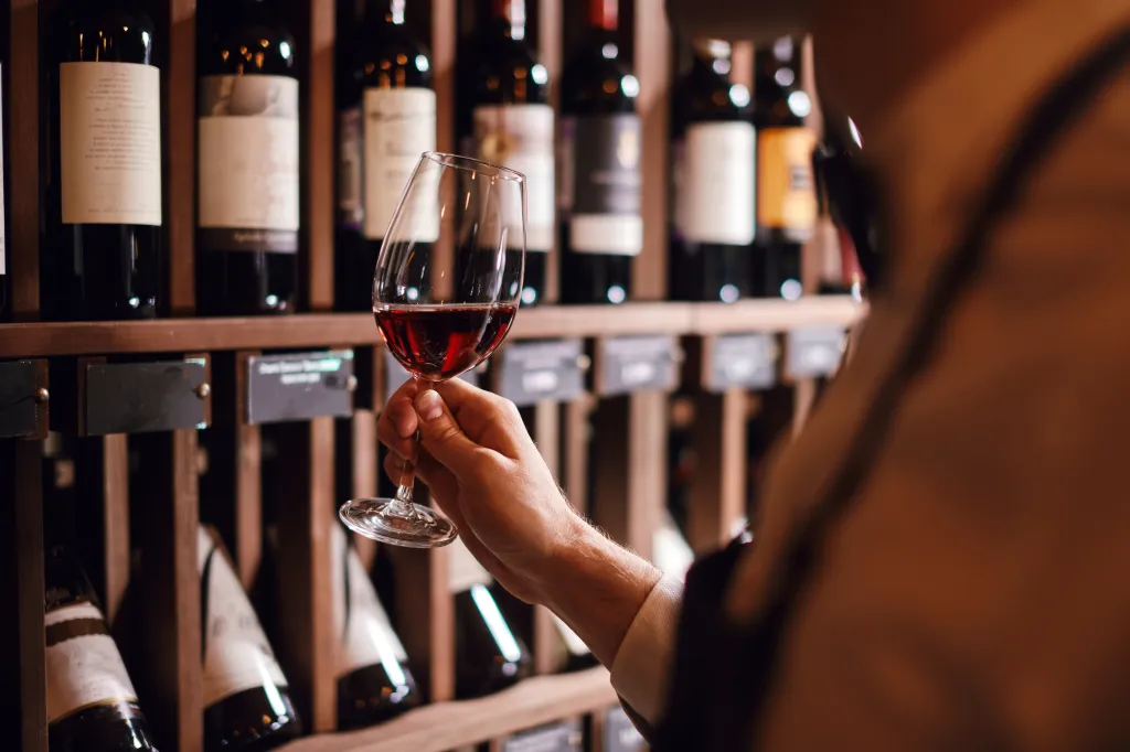 A man holding a glass of red wine, examining its tint, with bottles of wine on shelves in the background.