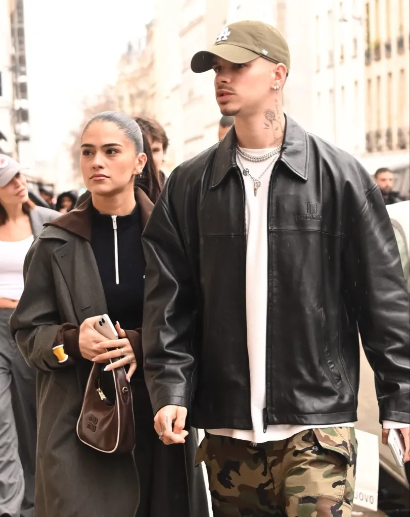 Romeo Beckham and Kim Turnbull walking together, Romeo in a black leather jacket and camouflage pants, and Kim in a dark coat carrying a small brown bag.