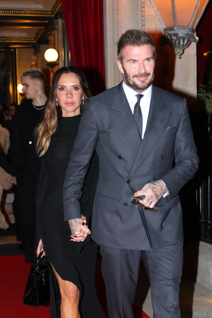 Victoria and David Beckham holding hands, with Victoria in a black dress and David in a gray double-breasted suit.