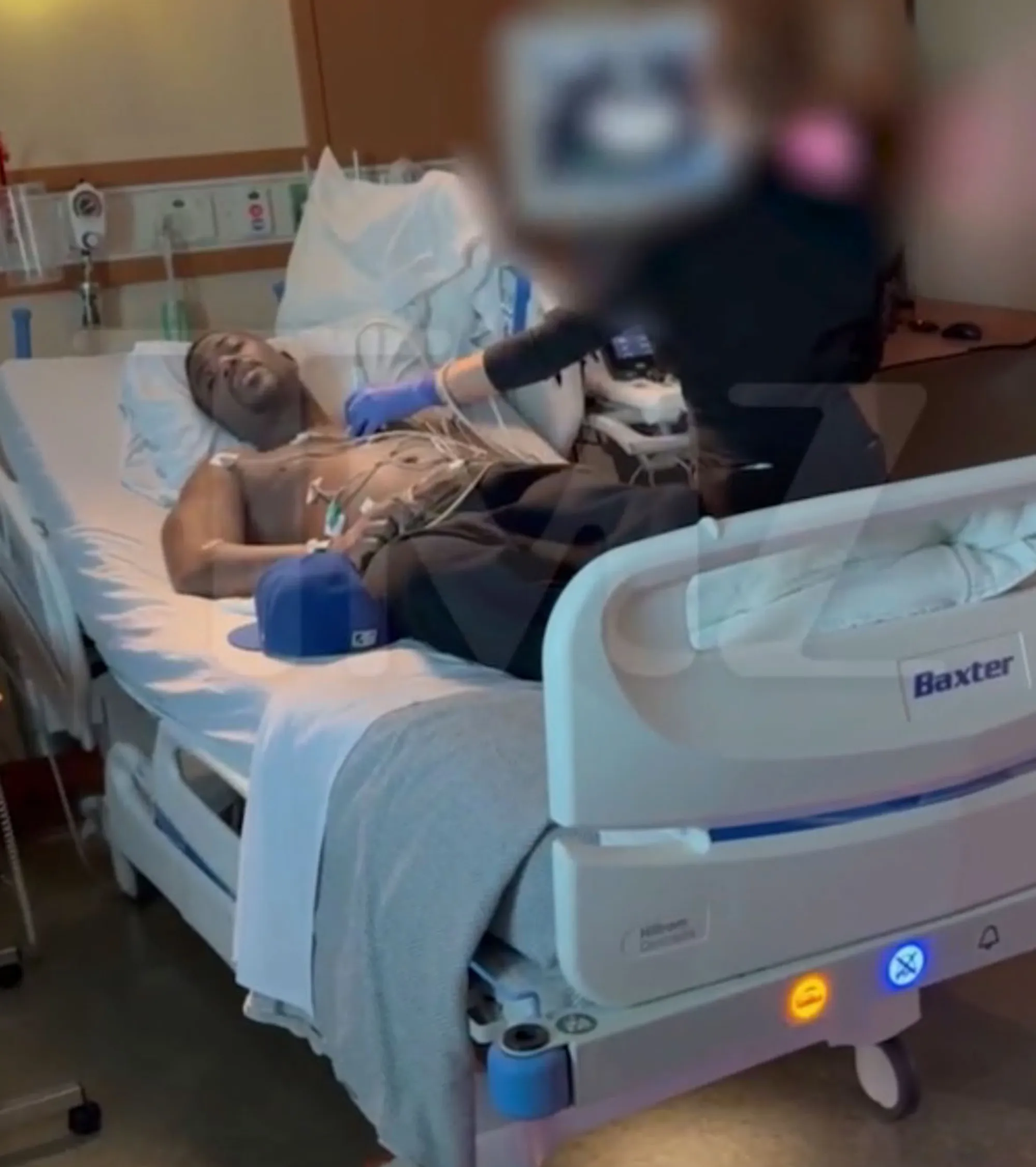 Ray J in a hospital bed with medical equipment attached, as a medical professional tends to him.