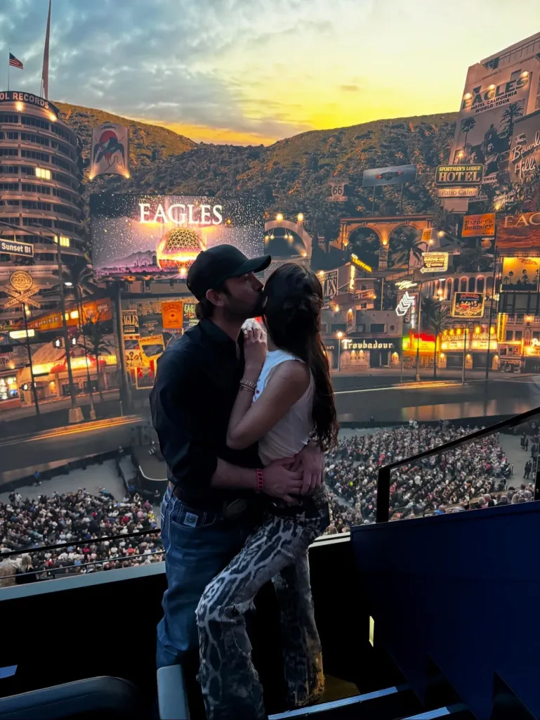 Bella Hadid and Adan Banuelos share a kiss at an Eagles concert, with the stage background featuring a Hollywood-themed cityscape.