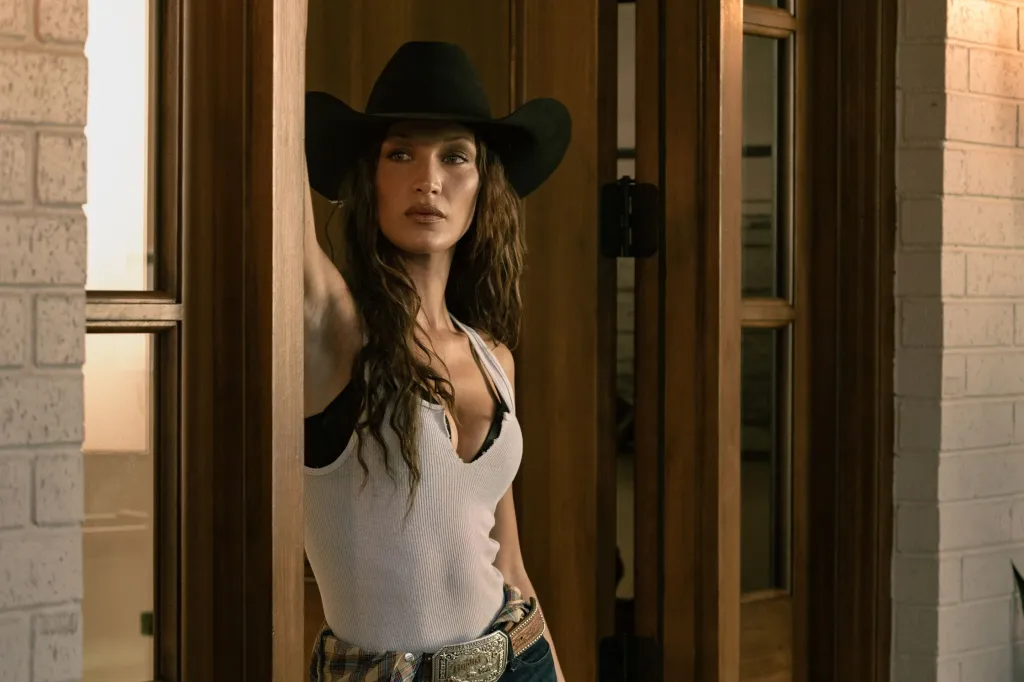 Bella Hadid in a white tank top and black cowboy hat leaning against a door frame.