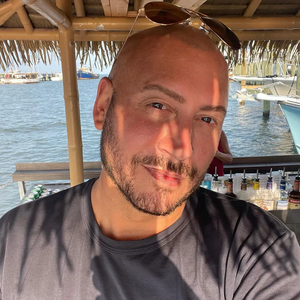 Jasen Kaplan smiling with sunglasses on his head in front of a body of water with boats.
