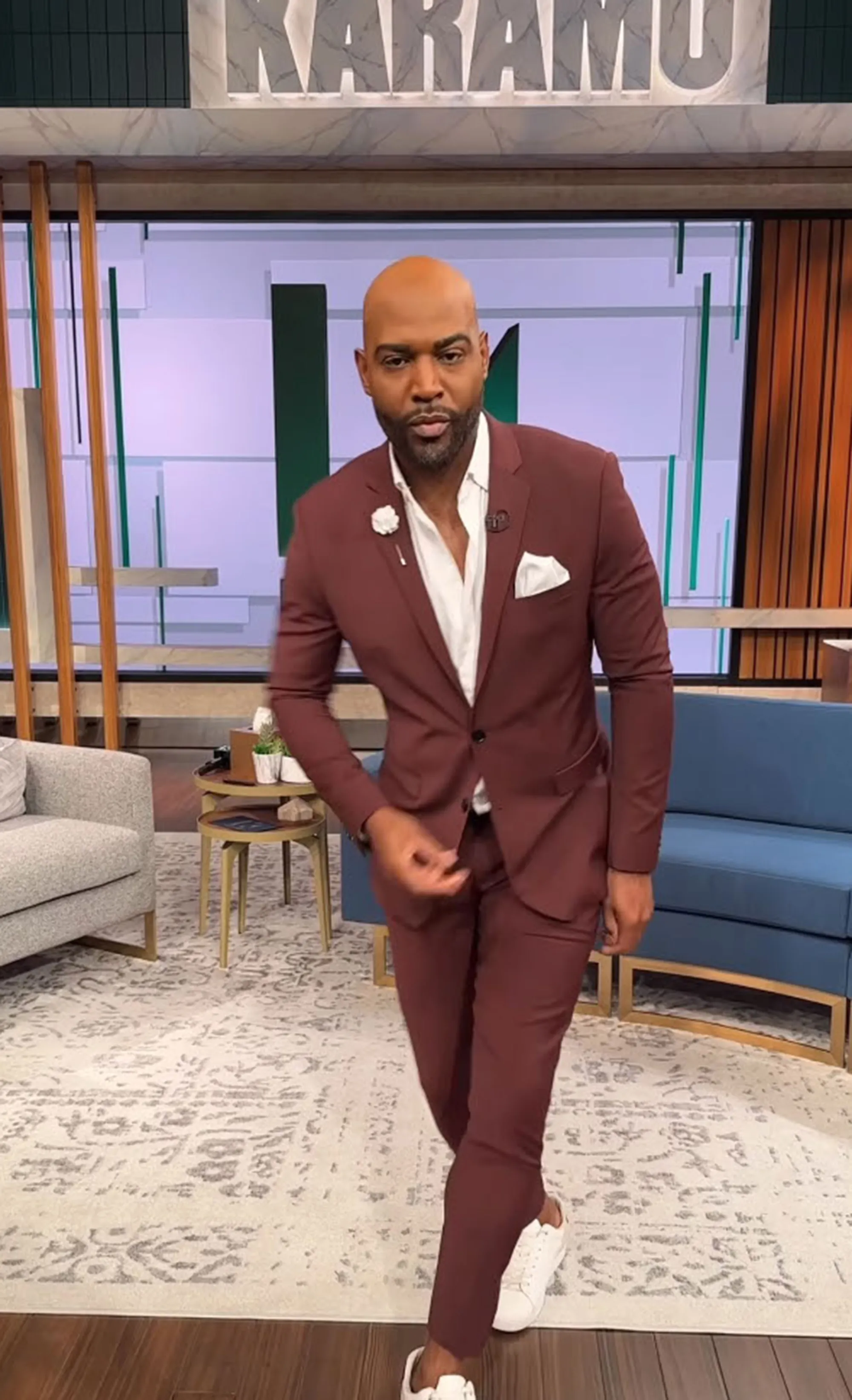 Karamo Brown in a maroon suit and white sneakers on a talk show set.