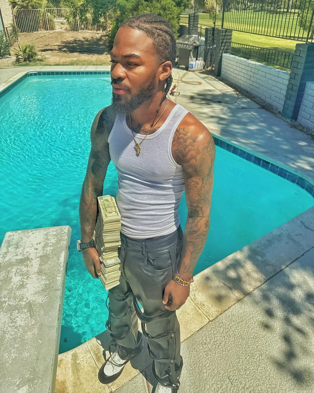 Twin Hector in a white tank top and black pants, holding a stack of cash next to a pool.