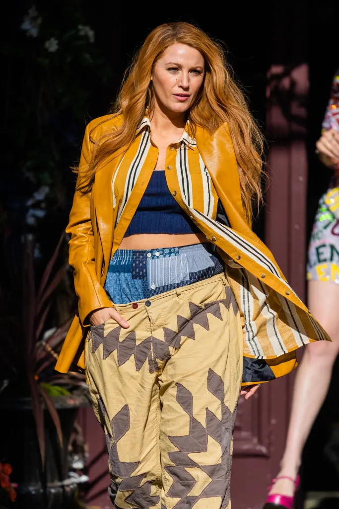 Blake Lively in character with long red hair, wearing a golden jacket over a striped shirt, blue crop top, and beige patterned pants.