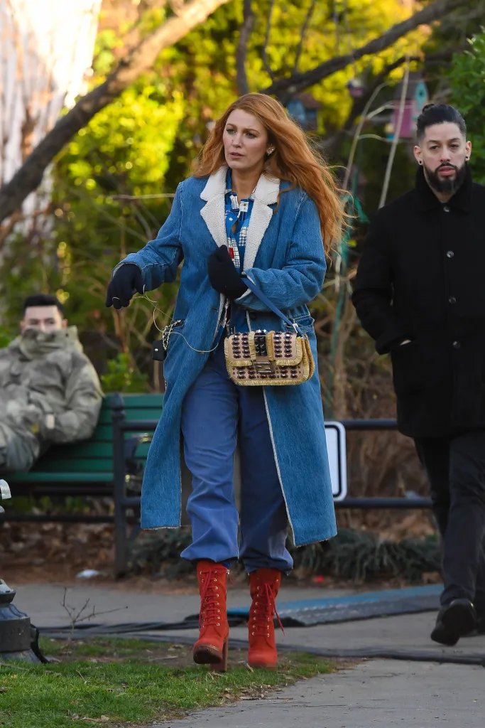 Blake Lively in a long denim coat with a cream shearling collar, blue pants, red boots, and carrying a fringed purse.