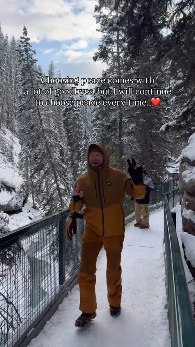 Bobby Berk in a mustard-colored winter coat and pants, making a peace sign on a snowy bridge surrounded by snow-covered pine trees, with the overlaid text, 