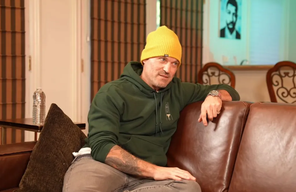 Brian Austin Green, wearing a yellow beanie and green hoodie, seated on a brown leather couch.
