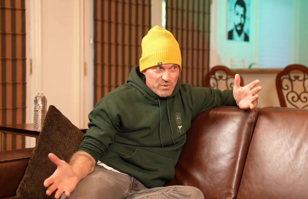 Brian Austin Green in a yellow beanie and green hoodie, seated on a brown leather couch and gesturing with his hands.