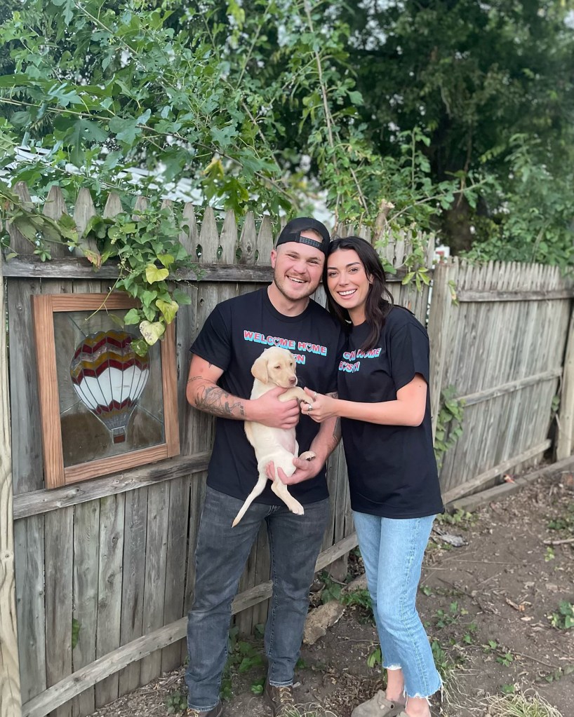 Brianna Chickenfry and Zach Bryan smiling and holding a yellow lab puppy in front of a fence.