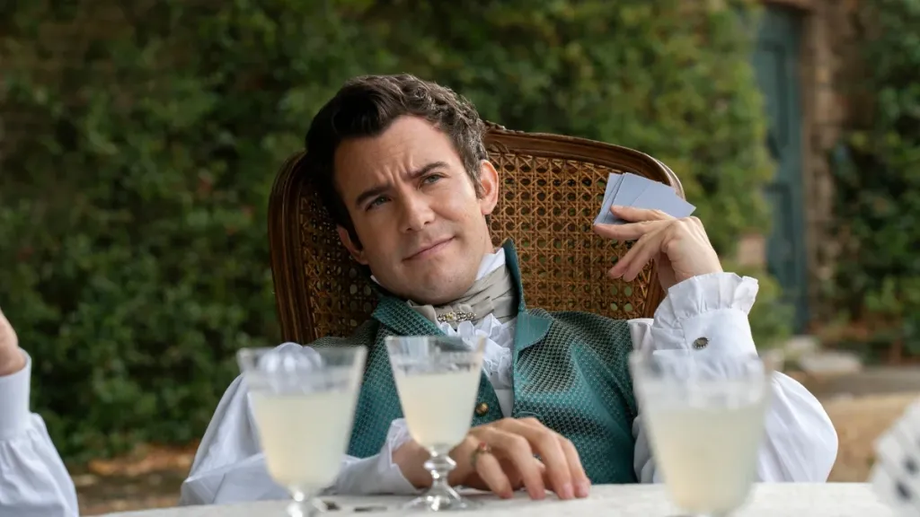 Benedict Bridgerton reclines in a wicker chair outdoors, holding playing cards with a relaxed, amused expression. He wears an ornate green waistcoat, white ruffled shirt, and cravat, with several pale drinks on the table in front of him with greenery behind him.