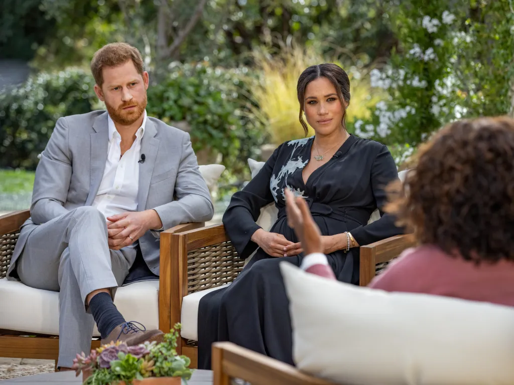Prince Harry, in a grey suit and white shirt, and Meghan Markle, in a black shirt with white pattern, being interviewed by Oprah Winfrey outdoors.