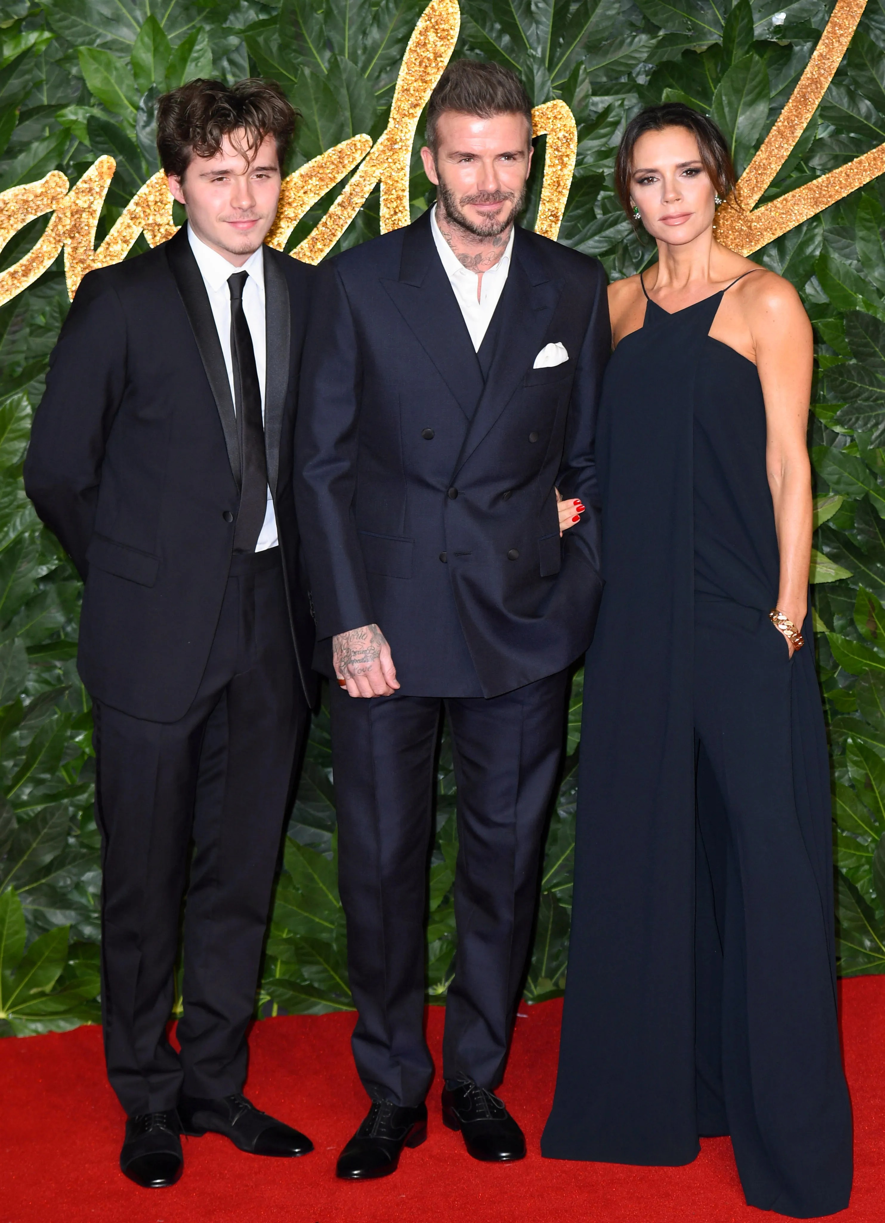 Brooklyn Beckham, David Beckham, and Victoria Beckham pose on a red carpet.