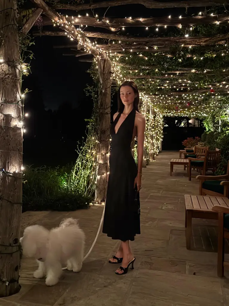 Nicola Peltz in a black dress standing on a patio next to a white dog, under a lit pergola at night.