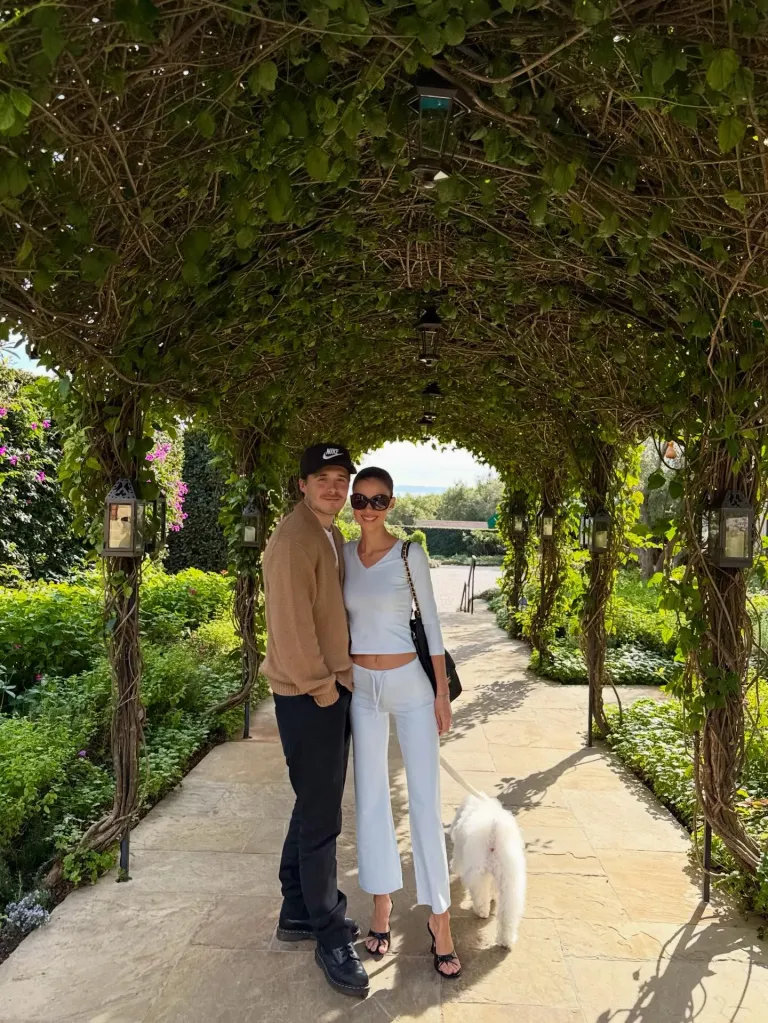 Brooklyn Beckham and Nicola Peltz standing under a vine-covered archway with their dog.