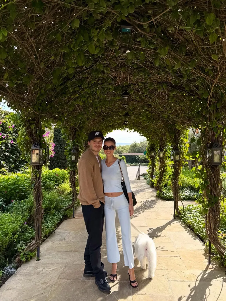 Brooklyn Beckham and Nicola Peltz standing under a leafy archway with their white dog.