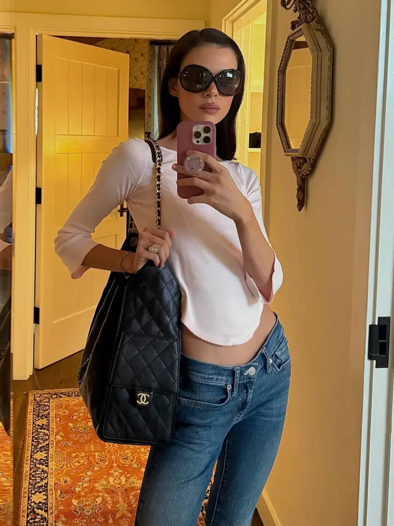 A woman in sunglasses and a pink top takes a mirror selfie while holding a Chanel bag.