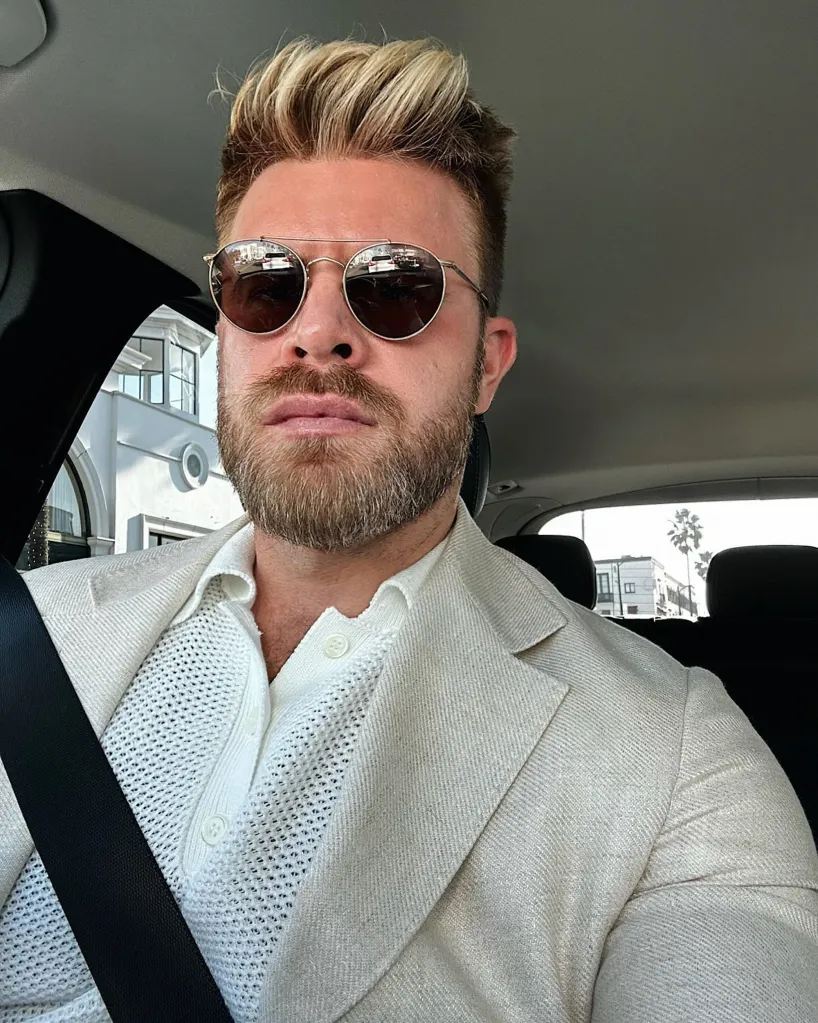 Man wearing sunglasses, a beige jacket, and a white knit shirt in a car.