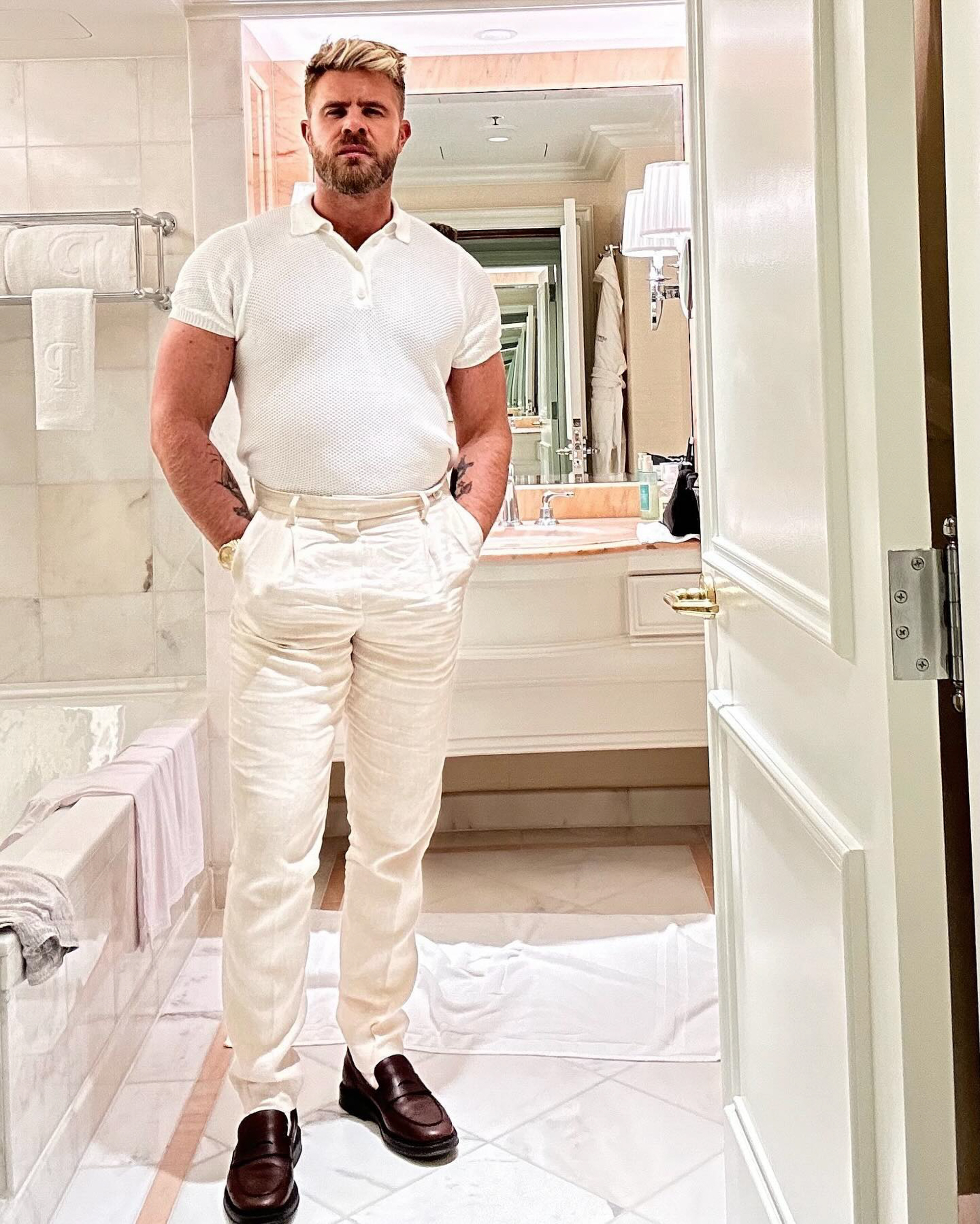 Hairstylist Justin Anderson standing in a white bathroom, hands in pockets, looking at the camera.