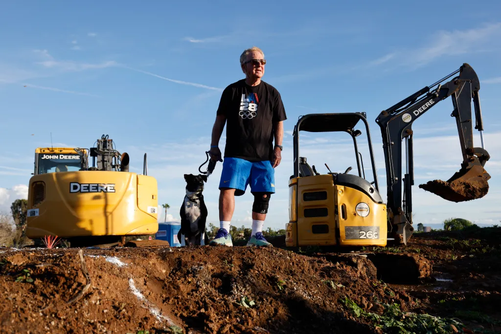 Bud Kling and his pit-boxer mix, Leia, standing on the remains of his home after the Palisades fire, with two John Deere excavators in the background.
