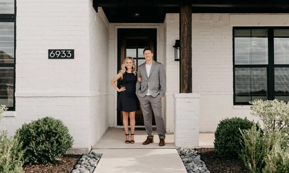 Two people, a woman and a man, standing on the porch of a white brick house with the number 6933.