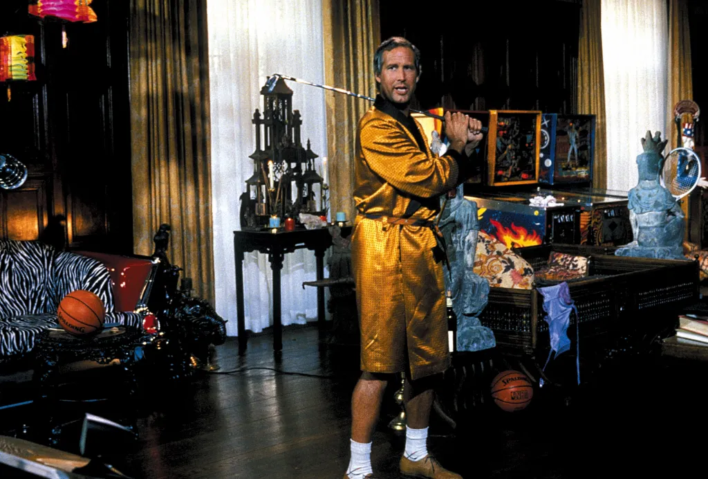 Chevy Chase in a gold bathrobe holding a golf club indoors.