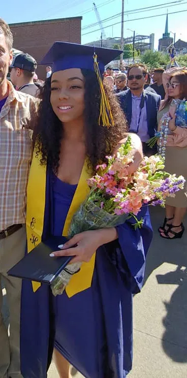 Carmen Anita Degregg at her high school graduation, wearing a royal blue gown and cap with a yellow stole, holding a bouquet of flowers and her diploma.