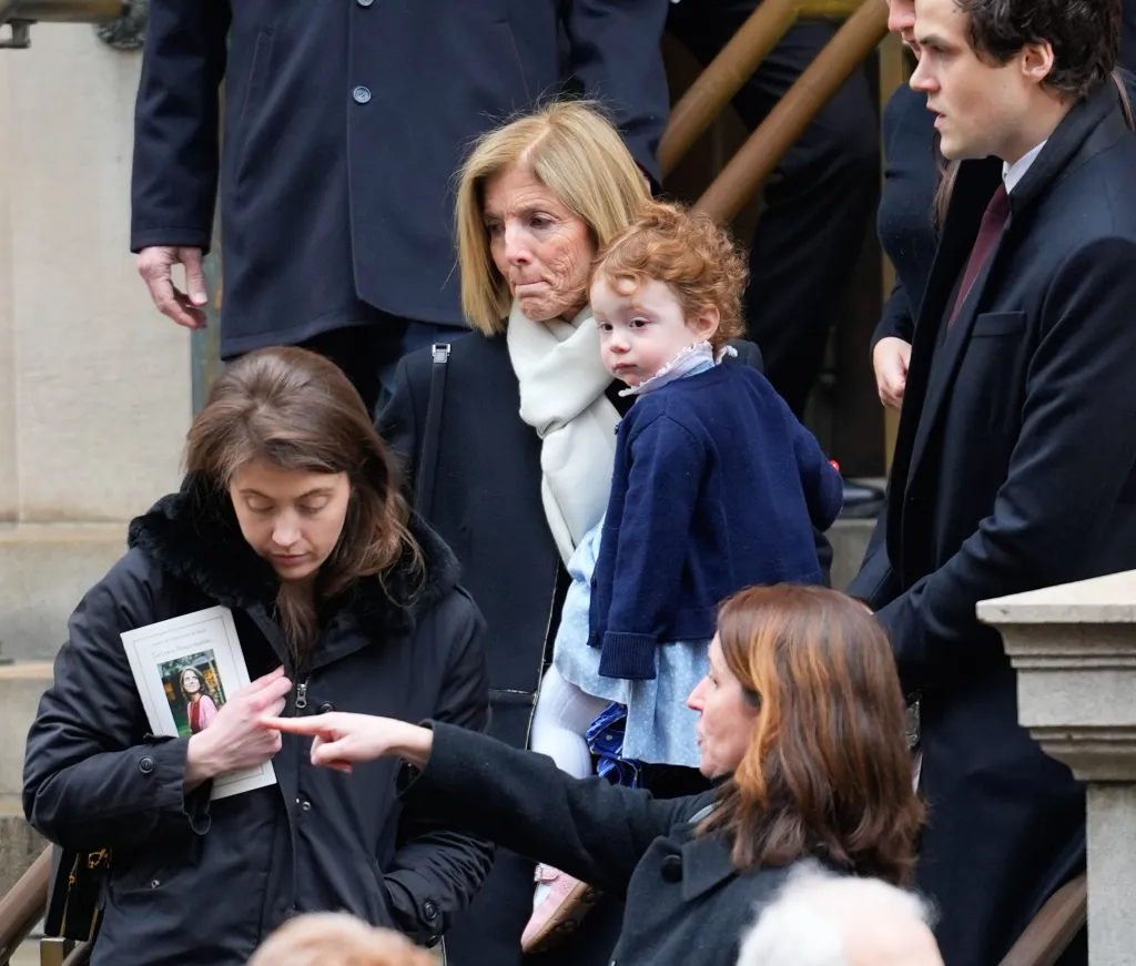 Caroline Kennedy-Schlossberg holding one of Tatiana's kids, with another woman holding a funeral program for Tatiana Schlossberg, and a young man beside them.
