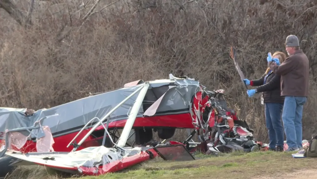 The wreckage of a red and grey plane after it crashed, with two people in the background examining a piece of the wreckage.