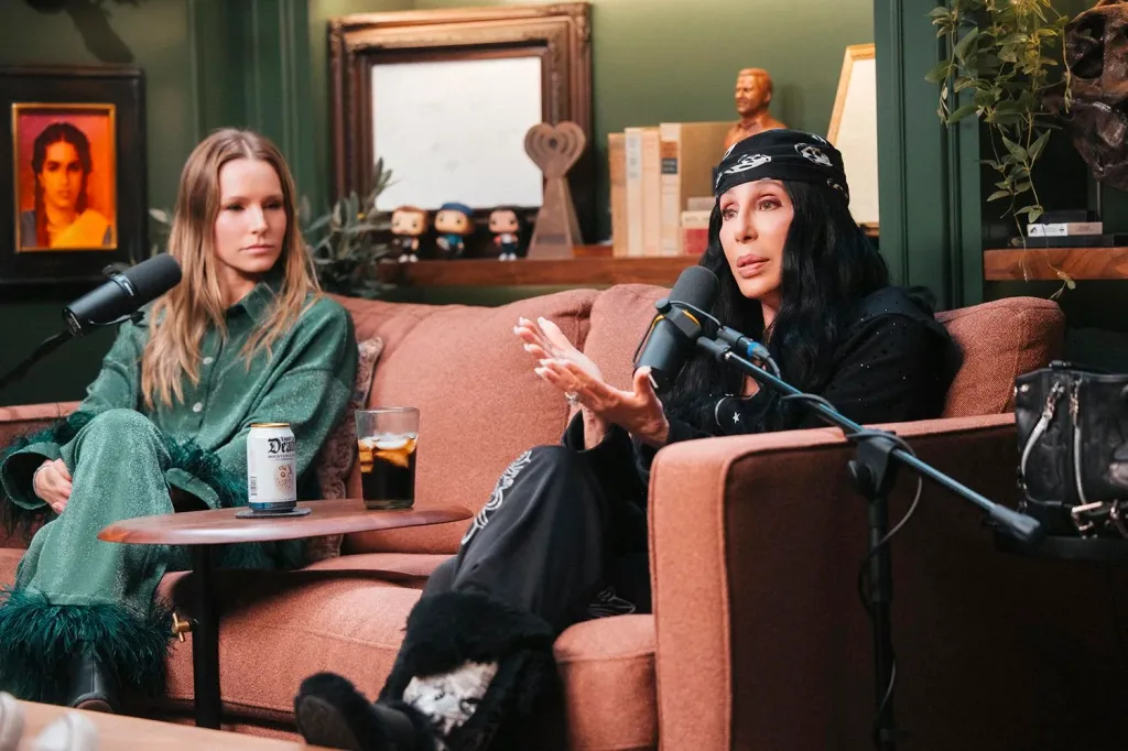 Kristen Bell and Cher sitting on a couch with microphones, engaged in conversation during a podcast.