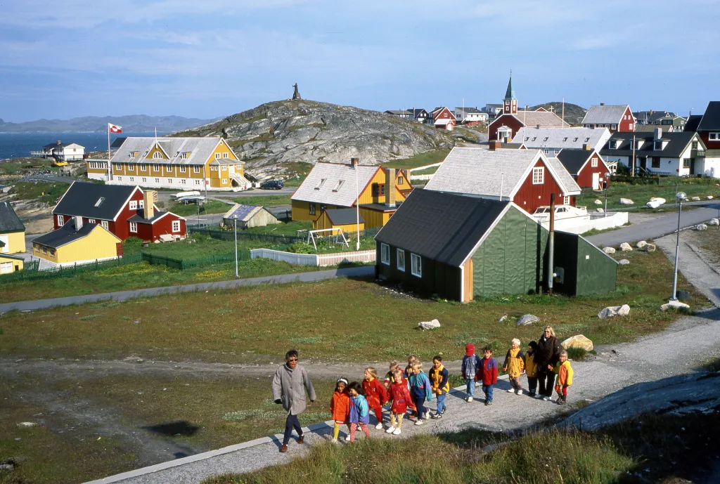 Children and teachers walk along a path in Nuuk, Greenland, with colorful houses, rocky hills, and the sea in the background.