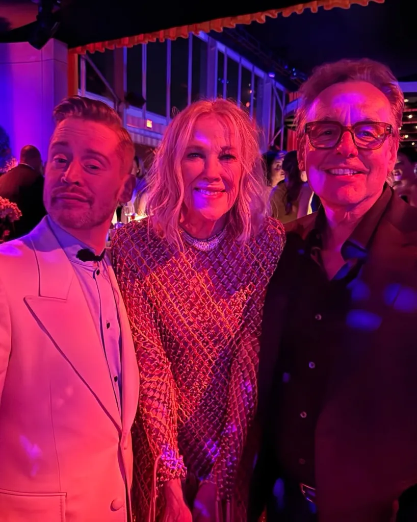A man in a white tuxedo, a woman in a metallic dress, and a man in a black suit smile for a photo at an event.