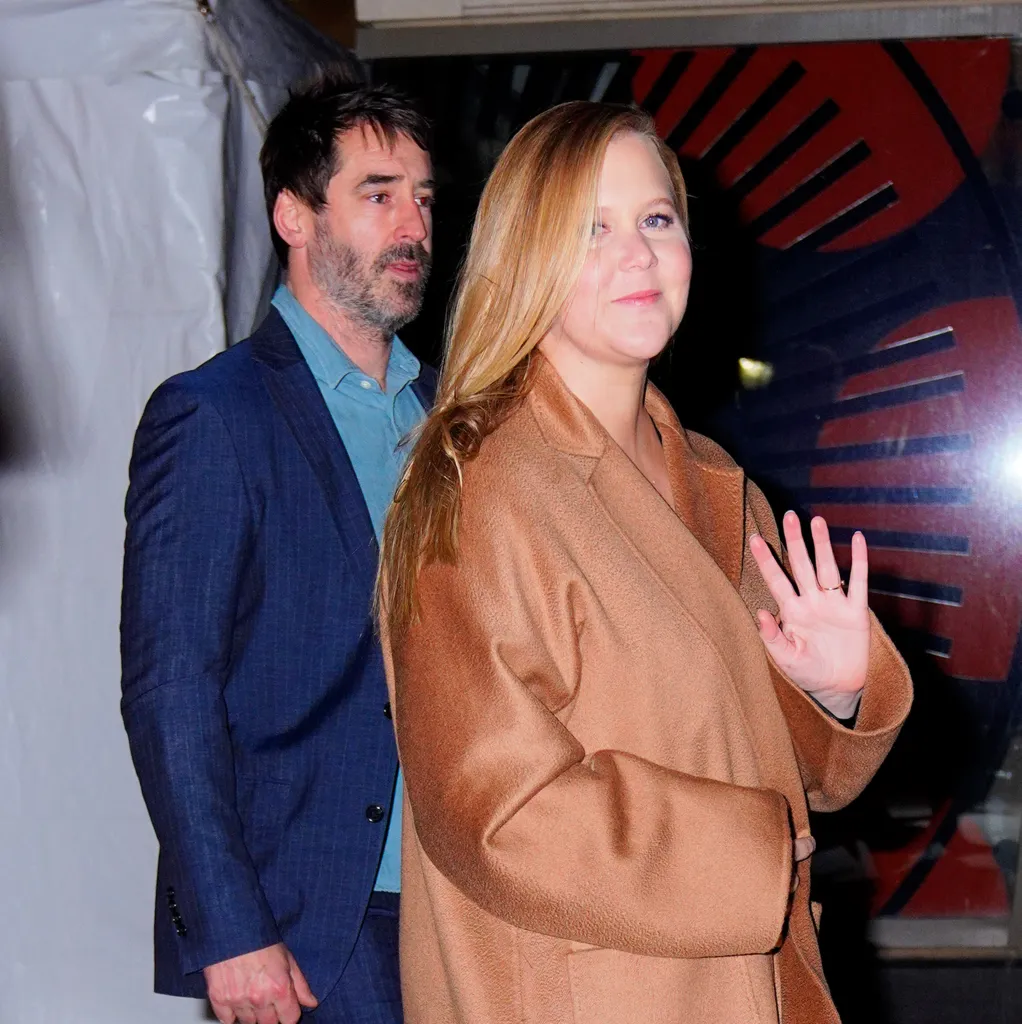 Amy Schumer in a brown coat waving, with Chris Fischer in a blue suit behind her.