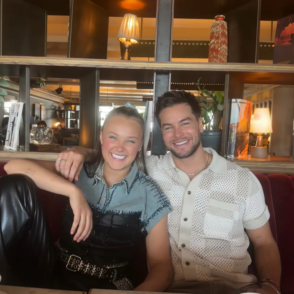 JoJo Siwa and Chris Hughes smiling while sitting together on a red couch.