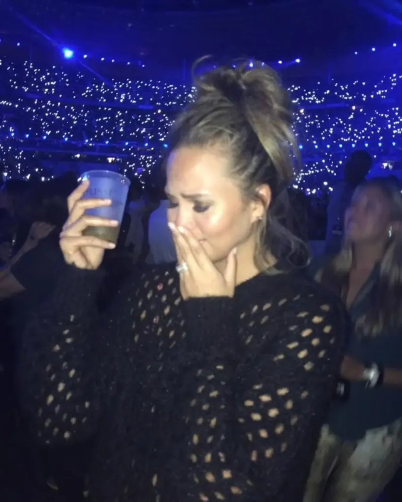 Chrissy Teigen holding a drink, with one hand covering her face, looking emotional at a concert with a crowd in the background.