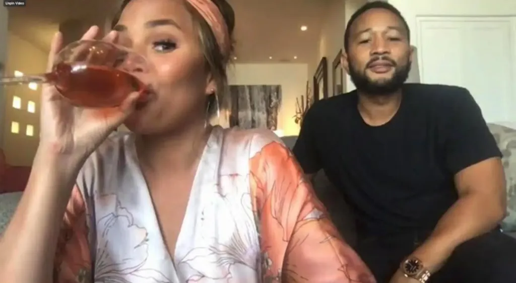 Chrissy Teigen drinking from a wine glass with John Legend next to her.