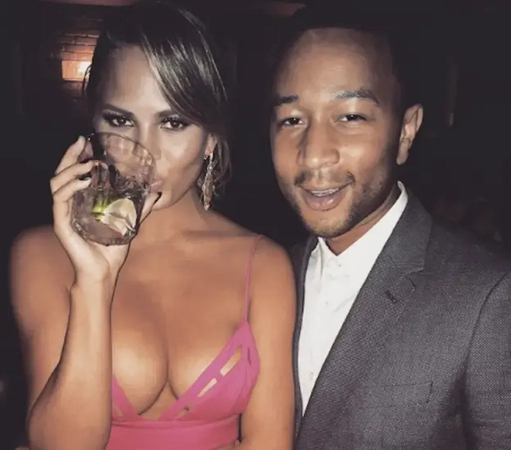 Chrissy Teigen drinking from a glass, with John Legend smiling beside her.